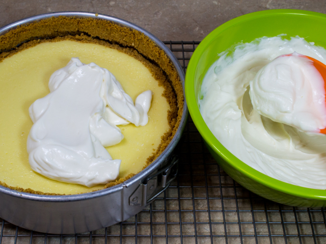 Sour cream mixture in green bowl being gently spooned over cheesecake filling.