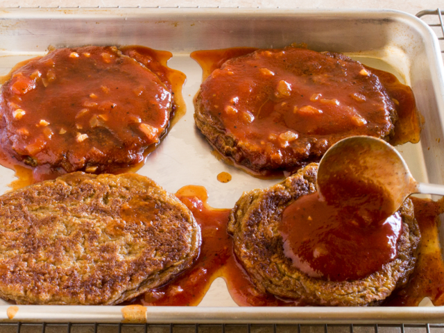 Four seitan steaks on a baking sheet and spooning barbecue sauce over each steak.