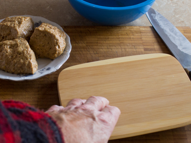 Three pieces of dough on a plate and one piece being pressed with a small cutting board.