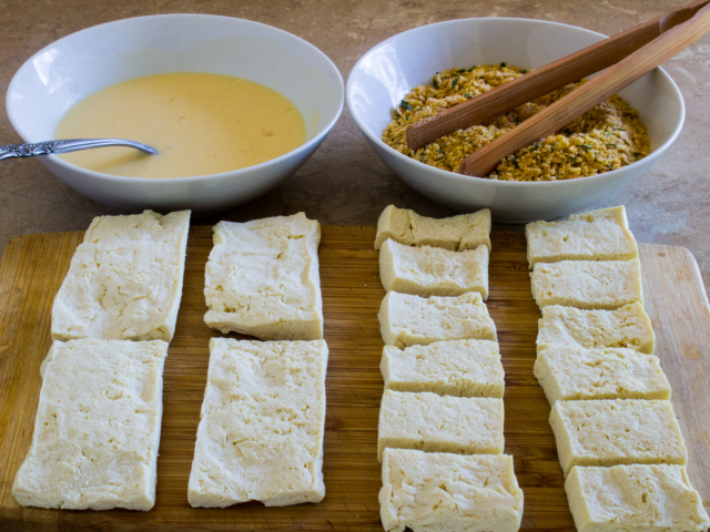 Bowl of egg batter, bowl of seasoned corn flake crumbs and thawed, sliced, tofu on cutting board.