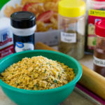 Green bowl of seasoned homemade corn flake crumbs with containers of herbs and spices