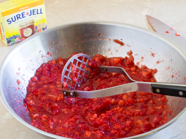 Chopped strawberries mashed in a large bowl with potato masher.